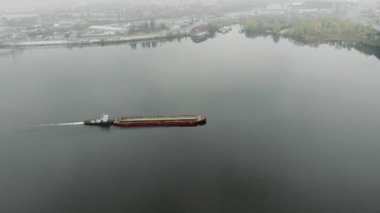 Büyük eski demir mavnası kumla dolu nehirde sanayi şehri boyunca sisli bir şekilde yol alıyor. Sanayileşmiş şehirde, gökyüzü manzaralı bir sis bulutunda yüzüyor.