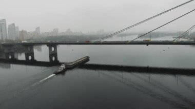 Endüstriyel kentteki beton köprü altında geniş nehir boyunca kum dolu büyük bir mavnayı taşıyan küçük bir römorkör. Şehir merkezinde yoğun bir duman içinde yüzen bir gemi.