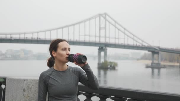 Retrato de mujer joven en forma deportiva bebiendo agua de la botella después de un duro entrenamiento. Atleta femenina descansando y bebiendo isotónica de la botella después de ejercicios de fitness al aire libre — Vídeo de stock