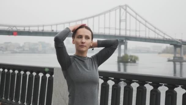 Donna legando i suoi corti capelli lisci in coda di cavallo, facendo acconciatura prima di allenamenti all'aperto duro allenamento e corsa, bellissimo paesaggio con ponte nella nebbia sullo sfondo — Video Stock