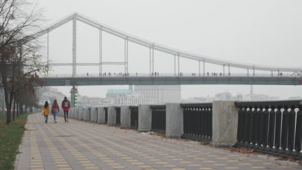 Menschen in heller Kleidung spazieren auf der Stadtpromenade entlang des Flusses mit schöner Brücke im Nebel im Hintergrund — Stockvideo