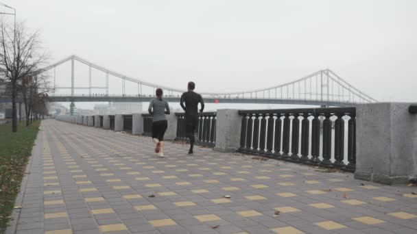 Běžící sportovní pár běhá po městské promenádě podél řeky. Fitness běžec muž a žena je těžký trénink pro každodenní cvičení. Pár mladých sportovců s krásnými těly běží na hubnutí — Stock video