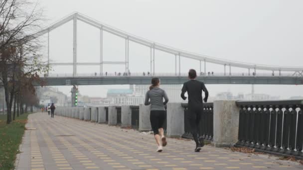 Motiverad passform smalt par är hård träning och gör utomhus träning. Kvinna och man intensiv kör längs staden kaj i dimma. Kvinnliga och manliga löpare som motionerar dagligen för viktminskning — Stockvideo