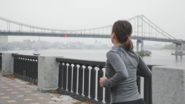 Jonge sportvrouw met prachtig mager lichaam loopt 's morgens in de mist langs de rivier. Vrouwelijke atleet in activewear is harde training en het doen van trainingen voor ultra marathon, brug op de achtergrond — Stockvideo