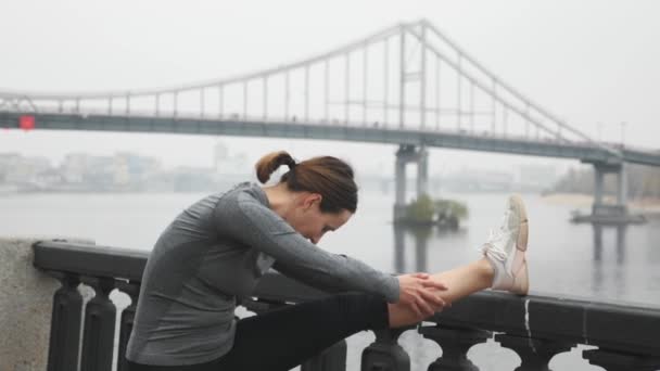 Piękna wysportowana kobieta rozciągająca się przed treningiem. Młoda biegaczka ćwiczy fitness w przemysłowym centrum miasta z mostem i mgłą na tle. Kobieta robi trening uliczny — Wideo stockowe