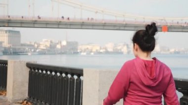 İskandinav yürüyüşü. Tombul güdümlü genç bir kadın şehir nehri gezintisinde sırıklarla yürüyor.
