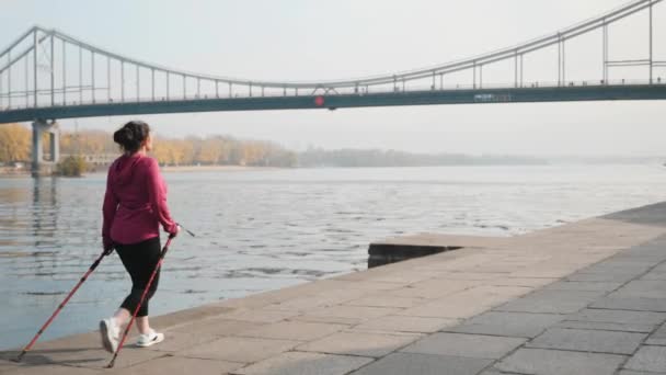 Подходит пухленькая женщина с палками для скандинавской ходьбы, тренирующаяся по утрам для соревнований и марафона. Концепция здоровья — стоковое видео