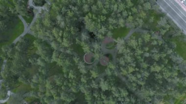 Büyük yeşil çam ağaçları, göl ve çocuk parkı olan güzel bir şehir parkı. Yaya yolları ve bankları olan park alanının havada dönen görüntüsü