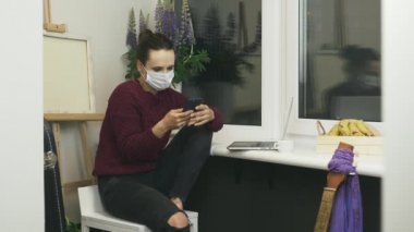 Koruyucu tıbbi maske takan genç esmer kadın akıllı telefon kullanıyor, dokunuyor ve ekranı kaydırıyor. Kadın elinde telefonla balkonda oturuyor, online haberleri izliyor, internette sohbet ediyor.