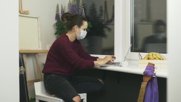Perempuan bertopeng wajah medis pelindung duduk di balkon apartemen modern dan bekerja pada komputer laptop jarak jauh. Wanita selesai bekerja pada notebook di tempat kerja rumah selama isolasi diri — Stok Video