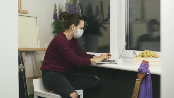 Mujer en la máscara protectora de la cara médica sentado en el balcón en el apartamento moderno y trabajando en el ordenador portátil de forma remota. Mujer terminó de trabajar en el portátil en el lugar de trabajo del hogar durante el auto-aislamiento — Vídeos de Stock