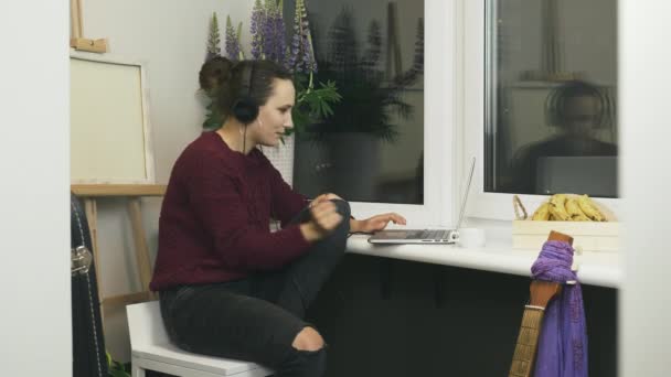 La joven mujer feliz se quita los auriculares mientras está sentada en el balcón y trabaja en la computadora portátil en el lugar de trabajo remoto en casa. Mujer freelancer haciendo trabajo a distancia. Estudiante estudiando en casa — Vídeos de Stock