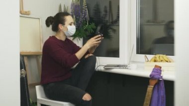 Koruyucu tıbbi maske takan genç bir kadın, rahat, modern bir dairede dizüstü bilgisayarda çalışırken kulaklık takıyor ve müzik dinliyor. Evden kendini soyutlama üzerine çalışmak