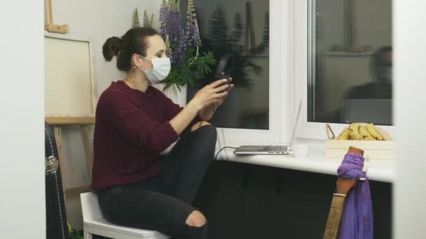 在舒适的现代公寓里，戴着防护口罩的年轻女性在远程工作场所的笔记本电脑上工作时，戴上耳机，听着音乐。在家里进行自我隔离工作 — 图库视频影像