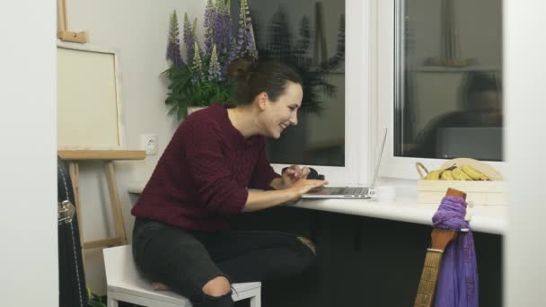快乐的黑发女人在舒适的现代阳台上一边笑一边看笔记本电脑上的在线新闻。女学生使用笔记本电脑，在社交媒体上与朋友聊天，大笑 — 图库视频影像