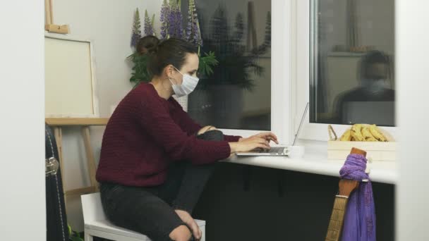 Młoda kobieta freelancer w masce ochronnej medycznej twarzy pracuje zdalnie nad nowym projektem pozostając w domu w przytulnym mieszkaniu. Kobieta pracująca na laptopie w odległym miejscu pracy. Kobieta korzystająca z laptopa w domu — Wideo stockowe