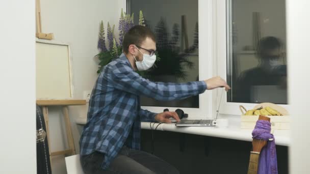戴防护医疗面罩的男人打开手提电脑，坐在舒适的现代公寓阳台上开始工作。男性自由职业者在家里写本关于自我隔离的笔记本 — 图库视频影像