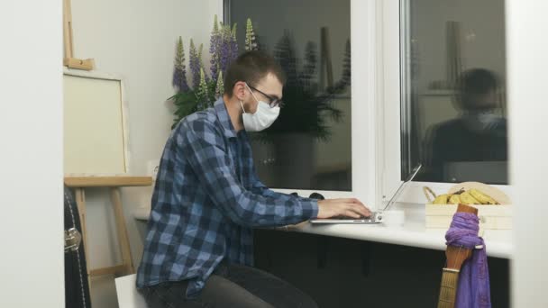 Muž na volné noze pracuje na vzdáleném projektu v domácím studiu na balkóně. Muž student v lékařské masce obličeje dělá on-line projekt na notebooku a studuje on-line na internetu zůstat doma — Stock video