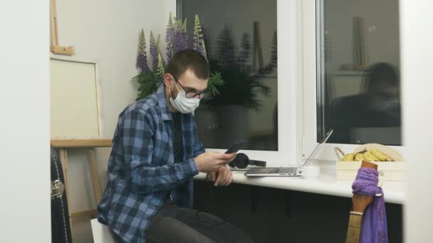 Jeune homme en lunettes et masque médical de protection utilisant un smartphone, bavarder en ligne dans les médias sociaux avec des amis, regarder des formations en ligne, faire défiler les nouvelles sur Internet assis sur le balcon à la maison — Video