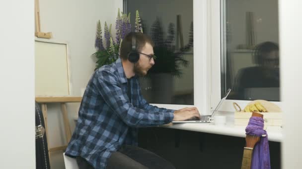 Junger Mann mit Kopfhörern, der Musik hört und auf der Tastatur seines Laptops tippt und aus der Ferne an einem neuen Projekt arbeitet. Männliche IT-Programmierer erstellen Codes für neue Arbeitsprojekte auf Notebook zu Hause — Stockvideo