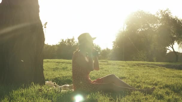 Fiatal nő visel retro szalma kalap és ruha ül kockás a parkban, és felveszi a fülhallgató, zenét hallgatni, és élvezi a gyönyörű nyári naplemente. A nő fülhallgatót vesz fel, miközben a parkban ül. — Stock videók