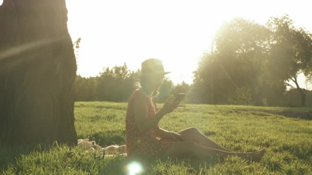 Dame souriante et heureuse écoutant la musique des écouteurs, regardant le coucher du soleil et profitant d'une chaude soirée d'été dans le parc. Femme appréciant week-end dans le parc public. Femme assise sur une prairie près d'un grand arbre — Video