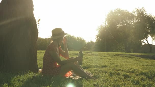 Mujer atractiva en sombrero de paja retro y vestido sentado en la hierba en el parque cerca de un gran árbol y disfrutar de la hermosa puesta de sol de verano. Linda chica sonriente bebiendo agua de la botella en el parque en un día soleado cálido — Vídeo de stock
