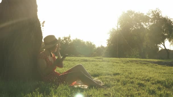 Улыбающаяся молодая женщина в соломенной шляпе и красном платье сидит в парке, опираясь на большое дерево и печатая сообщения, общаясь с друзьями в социальных сетях. Милая леди наслаждается прекрасным летним закатом в парке — стоковое видео