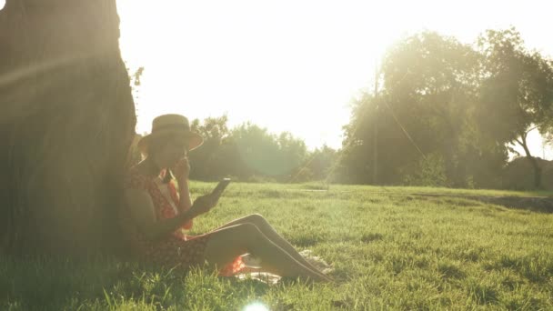 Jolie jeune fille en chapeau et robe rouge assis sur l'herbe dans le parc et met des écouteurs. Heureuse femme charmante écoutant la musique des écouteurs, assise dans le parc, penchée sur un grand arbre et profitant du coucher du soleil — Video