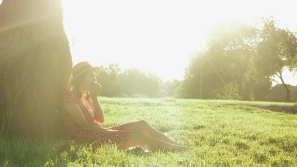 Vrouw zit in het park op gras op zonnige zomeravond. Jonge charmante dame met strohoed en jurk leunend op grote boom zittend op gras in het park, ontspannen en genieten van prachtige zonsondergang — Stockvideo