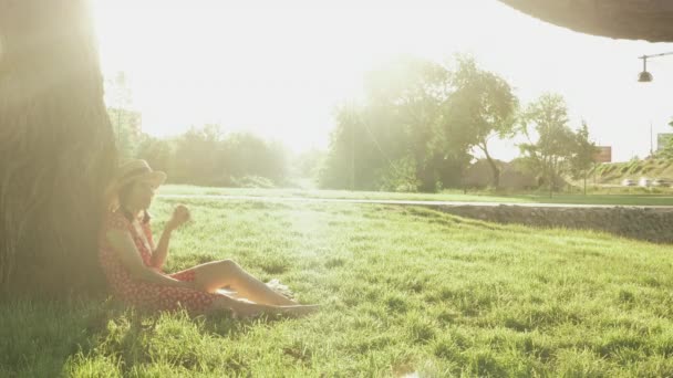 Charmante Dame in rotem Kleid und Strohhut sitzt auf Gras, lehnt an großen Baum im Park, ruht sich aus und isst Apfel. Frau mit Hut genießt Wochenende im Urlaub. Weiblich im Kleid entspannt — Stockvideo