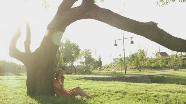 Счастливая молодая женщина отдыхает в парке возле большого дерева и ест яблоко. Милая женщина отдыхает в парке на закате. Очаровательная девушка в соломенной шляпе сидит на траве в парке и наслаждается летним вечером — стоковое видео