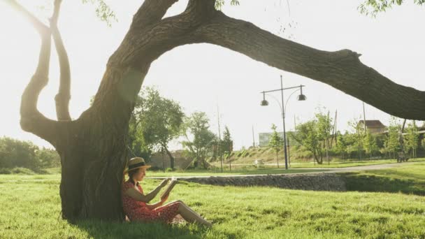 Kvinnan sitter i parken vid solnedgången och läser bok. Charmig kvinna sitter på gräs nära stora gröna träd i stadsparken och läsa bok på varm sommarkväll — Stockvideo