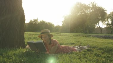 Güneşli yaz gününde çimenlikte kitap okuyan sevimli bir kadın. Kahverengi saçlı ve parkta edebiyat okuyan güzel, şık bir bayan. Kadın parkta dinleniyor ve günbatımının tadını çıkarıyor.