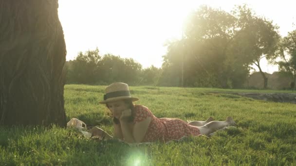 Morena mulher atraente em vestido relaxante no parque ao pôr do sol. Lady em chapéu deitado em xadrez no prado e diário de leitura, desfrutando de férias de fim de semana ao ar livre. Menina descansando no parque na noite ensolarada quente — Vídeo de Stock