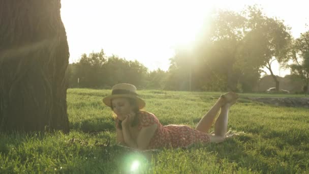 Jolie fille brune avec les jambes croisées allongées sur un plaid sur une prairie dans un parc public et un journal de lecture au coucher du soleil. Femme en chapeau et robe relaxant dans le parc et profiter d'une chaude journée ensoleillée en plein air — Video