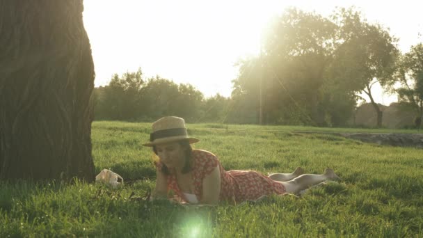 Dama de sombrero acostada en la hierba en el parque público y libro de lectura al atardecer. Mujer linda joven relajarse en el parque y disfrutar de las vacaciones de verano. Chica acostada en el prado y leyendo el diario del libro — Vídeo de stock