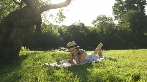 Mujer en sombrero acostada en el prado y leyendo libro en la naturaleza con el río en el fondo. Joven dama encantadora en vestido azul poco relajarse en la hierba en el parque cerca de gran árbol y disfrutar de las vacaciones de verano — Vídeo de stock