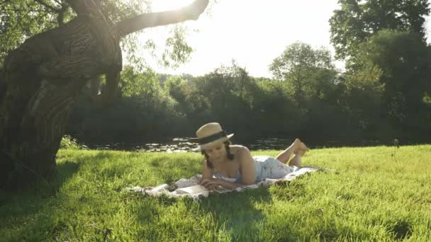 Fille reposant sur prairie par la rivière et profitant d'une chaude journée ensoleillée. Jolie femme brune en chapeau de paille couchée sur le plaid sur l'herbe sous un grand arbre près de la rivière, lire le journal du livre et se détendre en plein air — Video