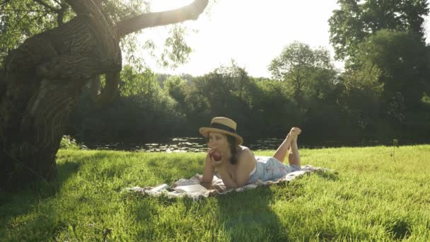 Young charming lady in hat and dress enjoying summer holidays at countryside. Cute woman lying on plaid on meadow under big tree by the river and eating apple. Female relaxing outdoor — Stock Video