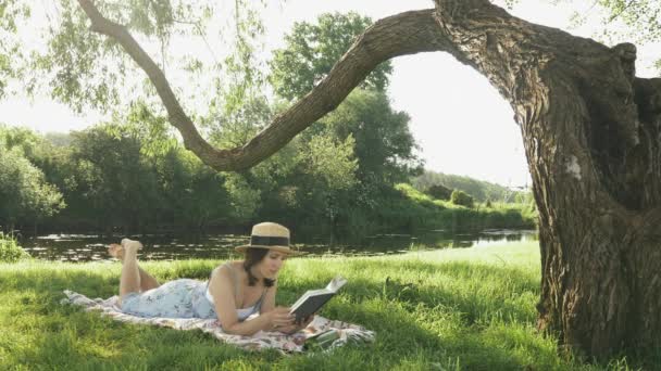 Joven dama encantadora en sombrero de paja y vestido acostado en la manta en el prado en el parque junto al río y libro de lectura. Linda chica morena relajándose en el parque y disfrutando del soleado clima de verano — Vídeo de stock