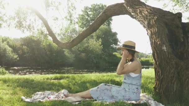 Charmante jeune femme assise sur plaid dans le parc au bord de la rivière et met des écouteurs. Fille profiter et écouter de la musique à partir d'écouteurs tout en se relaxant dans le parc sous un grand arbre au soleil journée d'été — Video