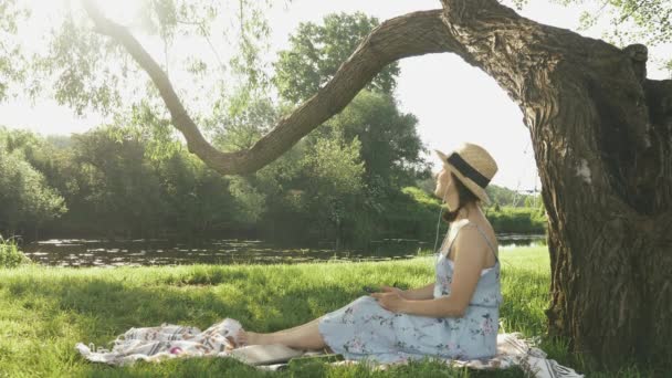 Joven mujer hermosa escuchando música de auriculares en el parque junto al río en el día soleado y cálido. Encantadora linda dama sentada en la hierba cerca de un gran árbol en el parque, escucha música y disfruta de las vacaciones de verano — Vídeo de stock