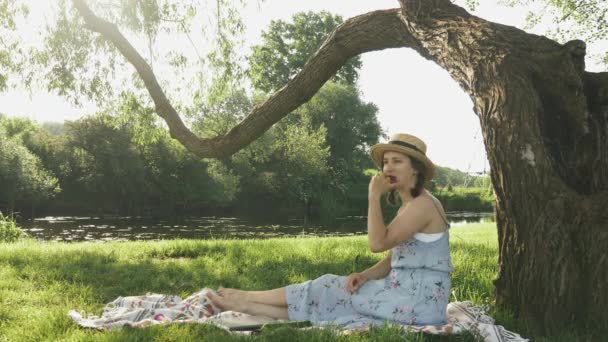 Спокойная очаровательная женщина в соломенной шляпе и платье едят яблоко, сидя на одеяле на траве в парке у реки. Леди, отдыхающая на улице и наслаждающаяся прекрасной природой в солнечный летний день — стоковое видео