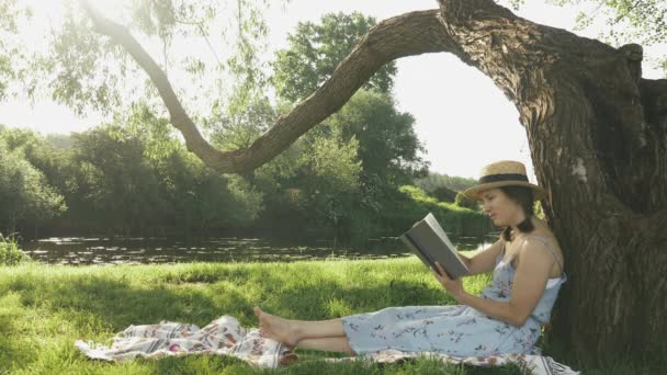 Dívka se opírá o velký strom, zatímco relaxuje v parku. Ženské studentky tráví čas v parku a čtou knihy. Mladá roztomilá dáma v klobouku a šaty sedí na trávě pod stromem a těší slunečný letní den — Stock video