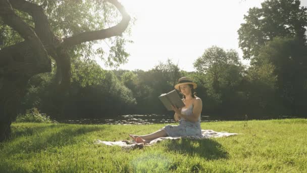 Joven mujer atractiva leyendo libro en el parque junto al río en el soleado día de verano. Encantadora linda dama relajándose en el parque el fin de semana, leyendo libro y disfrutando de las luces del sol. — Vídeos de Stock