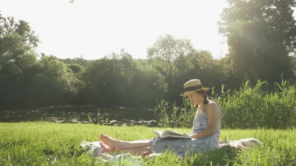 Mujer joven y serena disfrutando de un cálido día soleado en el parque junto al río. Linda mujer elegante sentada sobre una manta en la hierba cerca del río y leyendo el diario del libro. Señora relajándose al aire libre en el fin de semana de verano — Vídeos de Stock