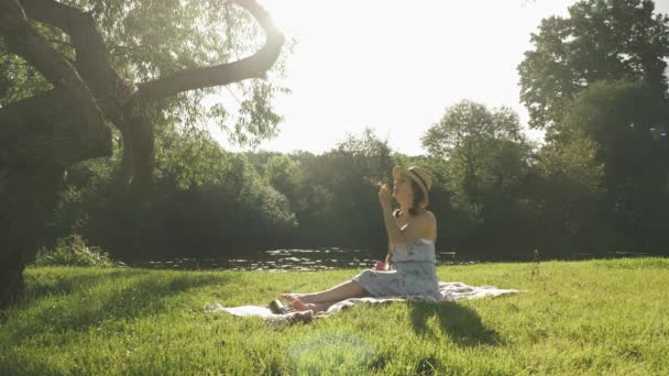 Stijlvolle vrolijke vrouw die bubbels blaast in het park bij de rivier. Jong brunette meisje zit op weide in de buurt van de rivier, plezier hebben, ontspannen en blaast bubbels op zonnige dag. Lady genieten van zomervakantie in park — Stockvideo