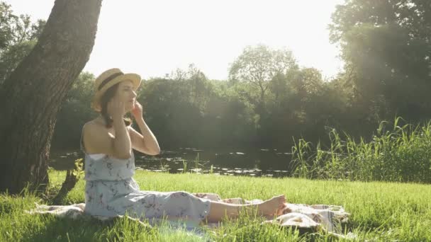 Chica con sombrero de paja y vestido sentado en la manta y se quita los auriculares, relajarse en el parque y disfrutar de un día soleado de verano. Linda dama feliz encantadora en fin de semana en el parque junto al río. Mujer descansando en el parque — Vídeo de stock