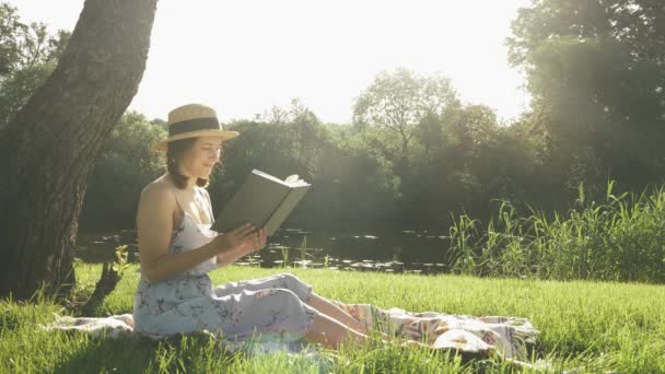 Gülümseyen mutlu kız şapkalı ve elbise giymiş kitap okuyor nehrin kenarındaki büyük ağacın altında çimlerin üzerinde oturuyor. Kadın tatilde hafta sonunun tadını çıkarıyor. Parkta ekose üzerinde oturan ve güneşli günün tadını çıkaran hoş bir bayan. — Stok video